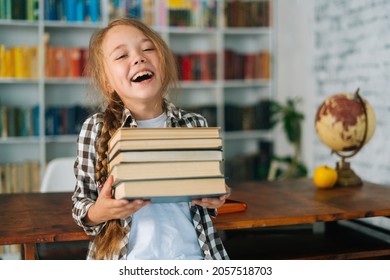 Medium Shot Portrait Of Laughing Elementary Child School Girl Holding Stack Of Books In Library At School, Looking At Camera. Cute Primary Pupil Schoolgirl Standing On Blurry Background Of Bookshelf.