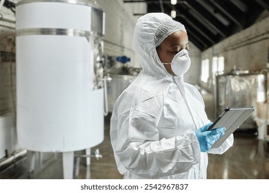 Medium shot of professional African American female technician in hazmat suit using tablet computer inspecting stainless tanks while controlling production process at food factory, copy space - Powered by Shutterstock