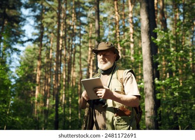 Medium shot of senior Caucasian male researcher standing in forest on sunny summer day and using digital tablet - Powered by Shutterstock