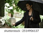 Medium shot of senior woman in mourning veil placing white lilies on stone of memorial monument at grave grieving over loss of loved one, while hiding from rain under black umbrella at cemetery