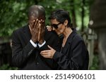 Medium shot of senior woman in sunglasses consoling crying husband at memorial ceremony. Multiethnic couple grieving loss of loved one at cemetery