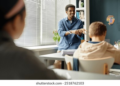 Medium shot of smiling African American male teacher talking to children in classroom with window blinds closed while counting on fingers explaining new topic to students at school, copy space - Powered by Shutterstock