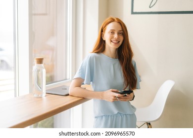 Medium shot of smiling charming young woman holding mobile phone, typing online messages standing by window in cozy light cafe. Pretty redhead Caucasian lady having leisure activity in coffee shop. - Shutterstock ID 2096965570