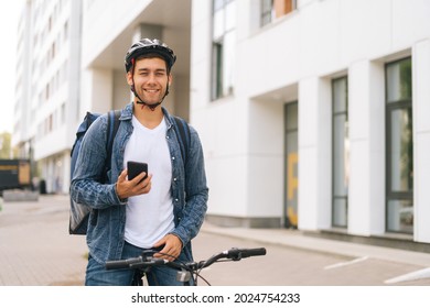 Medium shot of smiling handsome male courier with thermo backpack standing with bicycle in city street and using navigation app on phone. Delivery man looking for client address looking smartphone.