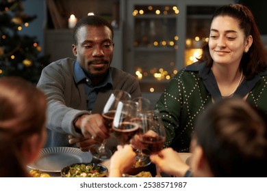 Medium shot of smiling Middle Eastern woman and Black man clinking glasses of red wine celebrating Christmas with friends during festive dinner at home party
