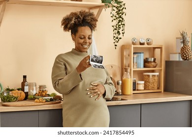 Medium shot of smiling pregnant Black woman holding ultrasound image of her baby while standing with hand on belly at kitchen counter with fresh vegetables - Powered by Shutterstock