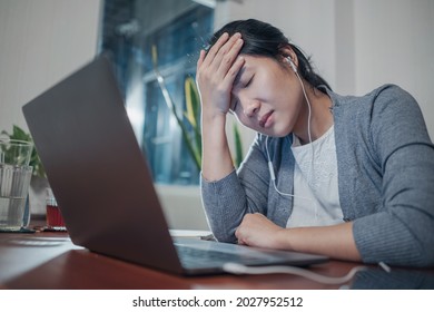 A medium shot of a stressed and frustrated young adult Asian woman, in a grey cardigan and white t-shirt inside working in home office with a laptop computer. head in hand. - Shutterstock ID 2027952512