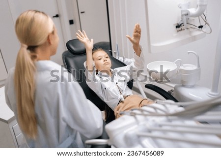 Medium shot of stressed little girl capricious and fooling sitting on dental chair and having dental treatment in dentistry clinic. Concept of children teeth treatment, pediatric checkup.