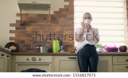 medium shot. Stylish bald woman drinking a hot drink while standing in the kitchen at home.