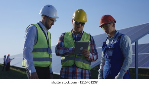 Medium shot of three electrical workers reviewing documents on a tablet during an inspection inbetween long rows of photovoltaic solar panels