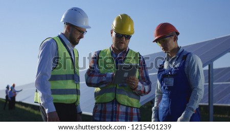 Medium shot of three electrical workers reviewing documents on a tablet during an inspection inbetween long rows of photovoltaic solar panels