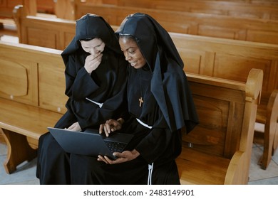 Medium shot of two ethnically diverse young and mature Catholic nuns sitting on pew enjoying surfing Internet on laptop