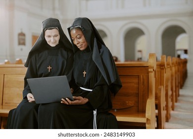Medium shot of two ethnically diverse young and mature nuns sitting on wooden bench in Catholic church working on laptop