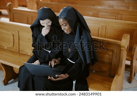 Medium shot of two ethnically diverse young and mature Catholic nuns sitting on pew enjoying surfing Internet on laptop