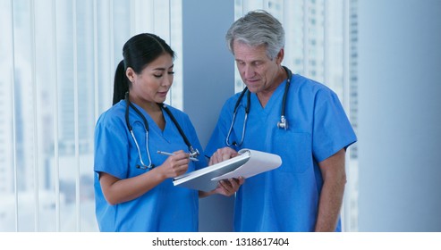 Medium Shot Of Two Friendly Medical Professionals Wearing Blue Scrubs Working Together. Japanese Woman Doctor And Caucasian Male Nurse Going Over Paperwork In Hospital