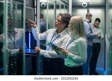 Medium shot of two women working in a data center with rows of server racks and checking the equipment and discussing their work - Powered by Shutterstock