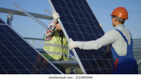 Medium Shot Of Two Workers In Safety Vests And Hardhats Installing Photovoltaic Panels On A Metal Basis At A Solar Farm