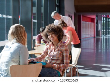 A Medium Shot Of Two Young Girls Talking To Each Other In School Hallway With Teacher Helping Students In Background.