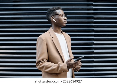 Medium shot of young African American businessman holding smartphone listening to voice message via earphones while standing outdoors - Powered by Shutterstock