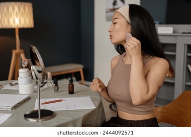 Medium shot of young Asian woman sitting at table looking into mirror doing gua sha facial massage - Powered by Shutterstock