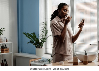 Medium shot of young Black woman brushing teeth engaged in text messaging on phone during morning self care routine in bathroom, copy space - Powered by Shutterstock