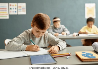 Medium shot of young boy focused on writing task in copybook sitting at desk while studying in classroom with blue wall at school, copy space - Powered by Shutterstock