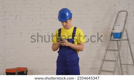 Medium shot of a young construction worker standing in the room, holding a smartphone, waiting for the answer. The man looks intemperate and concerned.