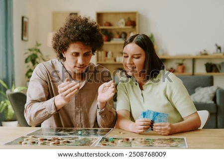 Medium shot of young ethnically diverse couple throwing dice while playing modern tabletop game in living room at home