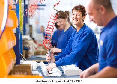 A medium shot of a young female worker smiling at camera while working on an aluminum light fittings on the production line with other workers. - Shutterstock ID 1847350087