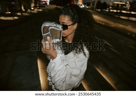 Medium shot of young Middle Eastern female celebrity hiding face with silver purse from paparazzi while walking through city with night lights in blurred background, copy space