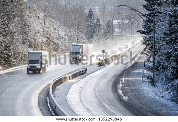 Medium size and Power capability day cab rig semi\
Truck for local deliveries with roof spoiler and dry van semi\
trailer running on the winter snowy and icy slippery road with\
divided concrete barrier