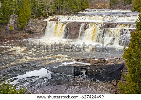 Medium size waterfall with a net in the river below. The net has ropes holding the posts to which it is attached. Spray from the waterfall creates a haze in the air. Trees line the river.