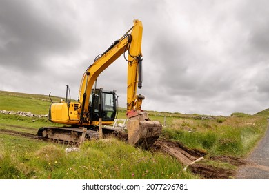 Medium Size Yellow Excavator Working In A Green Field. Building Family House Or Farm In A Country Side Concept. Dramatic Cloudy Sky In The Background. Heavy Machinery Equipment On A Job.