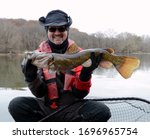 A medium sized brown  colored flathead catfish fish being held horizontally by a smiling man in a dry suit over a net on a river