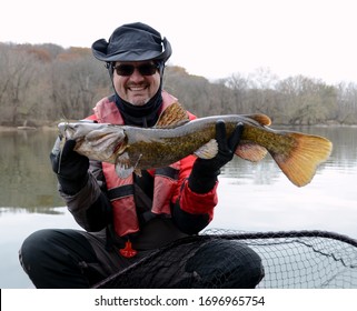 A medium sized brown  colored flathead catfish fish being held horizontally by a smiling man in a dry suit over a net on a river
