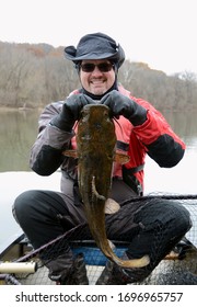 A medium sized brown olive colored flathead catfish fish being held vertically by a smiling man in a dry suit in a canoe on a river