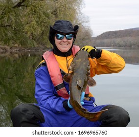 A medium sized brown olive colored flathead catfish fish being held vertically by a smiling woman in a dry suit in autumn