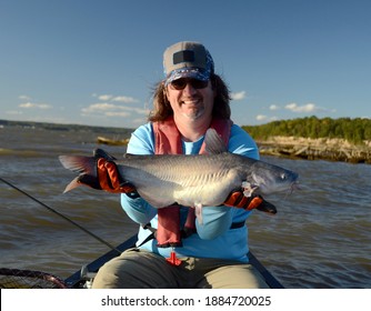A Medium Sized Silver Gray Blue Catfish Fish Being Held Horizontally In By A Smiling Man In A Canoe On A Rough River