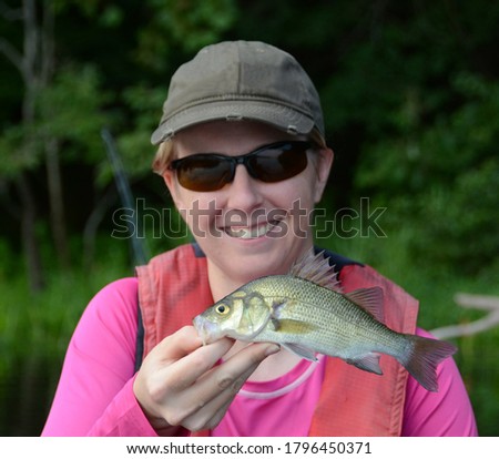 A medium sized white perch being held horizontally in one hand by a smiling woman in a baseball cap and pink shirt on a cloudy day on a river