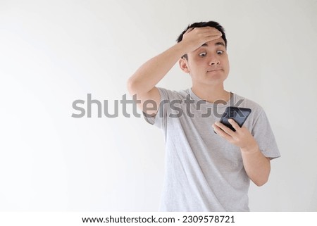 Medium and soft-focused shot of a young Asian man who wears a grey t-shirt and is facepalm while holding and looking at a smartphone. Shallow depth-of-field. Isolated white background.