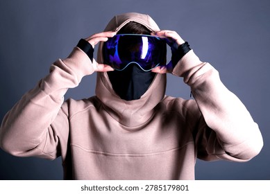 Medium studio shot of an unrecognizable winter sports person or model against a grey background. The subject is wearing a heavy pastel beige (nude color) oversized hoodie with the hood pulled up, a black protective face mask covering the face, and holding reflective blue snowboard goggles with both hands. Perfect for activewear catalog, protective gear presentation, winter fashion retail, and athletic clothing mockups.