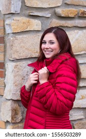 Medium Vertical Photo Of Pretty Young Woman With Long Mahogany Hair Smiling Seductively And Wearing Red Dress And Puffy Winter Coat While Standing In Front Of Stone And Brick House