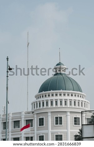 Medium view of a classical building featuring a prominent green dome roof, a waving red and white Indonesian flag on a flagpole, and a tall telecommunication antenna mast against a clear sky.