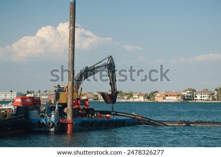 Medium Wide view of Looking out to Pass-a-grille channel. Dredge boat barge, crane and pipes in blue water. Blue sky white clouds and buildings in the distance. Near sunset with. At St. Pete Beach, FL