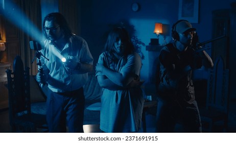 Medium-full photo capturing two ghost hunters and a victim going around the room in search for the cause of a paranormal activity, using EMF detector and filming it.