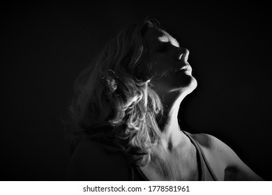 Meerkerk, The Netherlands- July 17 2020 : Black And White Low Key Portrait Of A Woman With Long Blond Hair Looking Up In The Air With Light Falling Along The Side Of Her Face And Dark Black Background