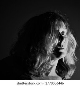 Meerkerk, The Netherlands- July 17 2020: Black And White Low Key Portrait From A Woman With Blond Hair Closing Her Eyes Black Background And Bright Light On Hair And Face