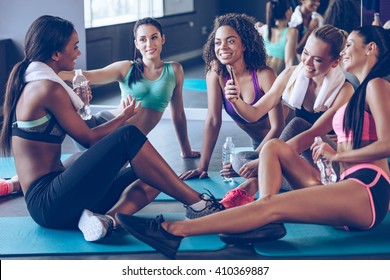 Meet Up On The Mat. Beautiful Young Women In Sportswear Discussing Something With Smile And Using Smartphone While Sitting On Exercise Mat At Gym