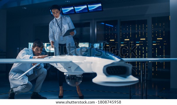 Meeting of Aerospace Engineers Working On\
Unmanned Aerial Vehicle / Drone Prototype. Aviation Scientists in\
White Coats Talking. Commercial Aerial Surveillance Aircraft in\
Industrial Laboratory