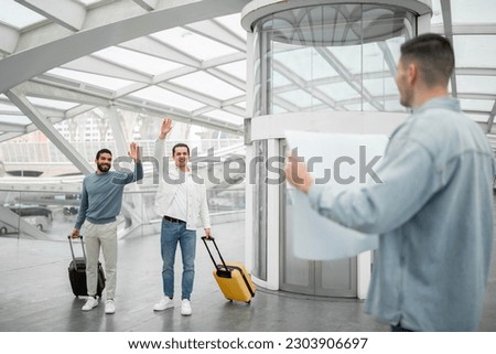 Meeting At Airport. Two Tourists Men Waving Hello To Guy With A Paper Sign Arriving At Terminal Indoor, Traveling On Business Or Vacation. Meet And Greet Service Offer Concept. Selective Focus